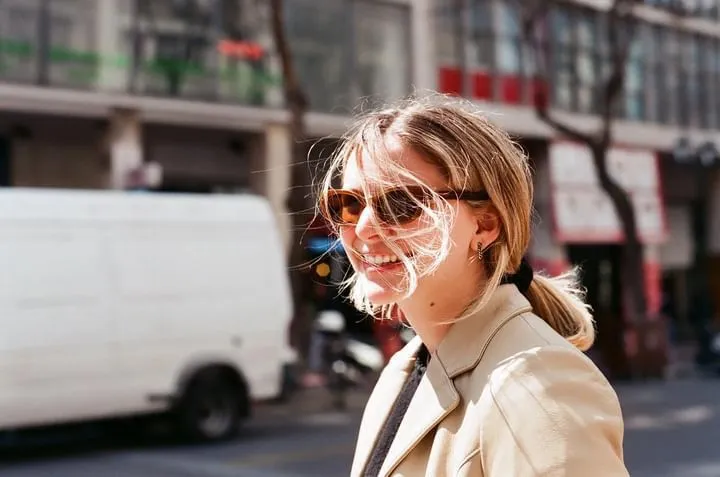 A photo of Hannah Kofman standing on a busy street in Athens. The background is blurred, Hannah is smiling. She's wearing a vintage beige leather jacket, sunglasses and has her hair in a ponytail.